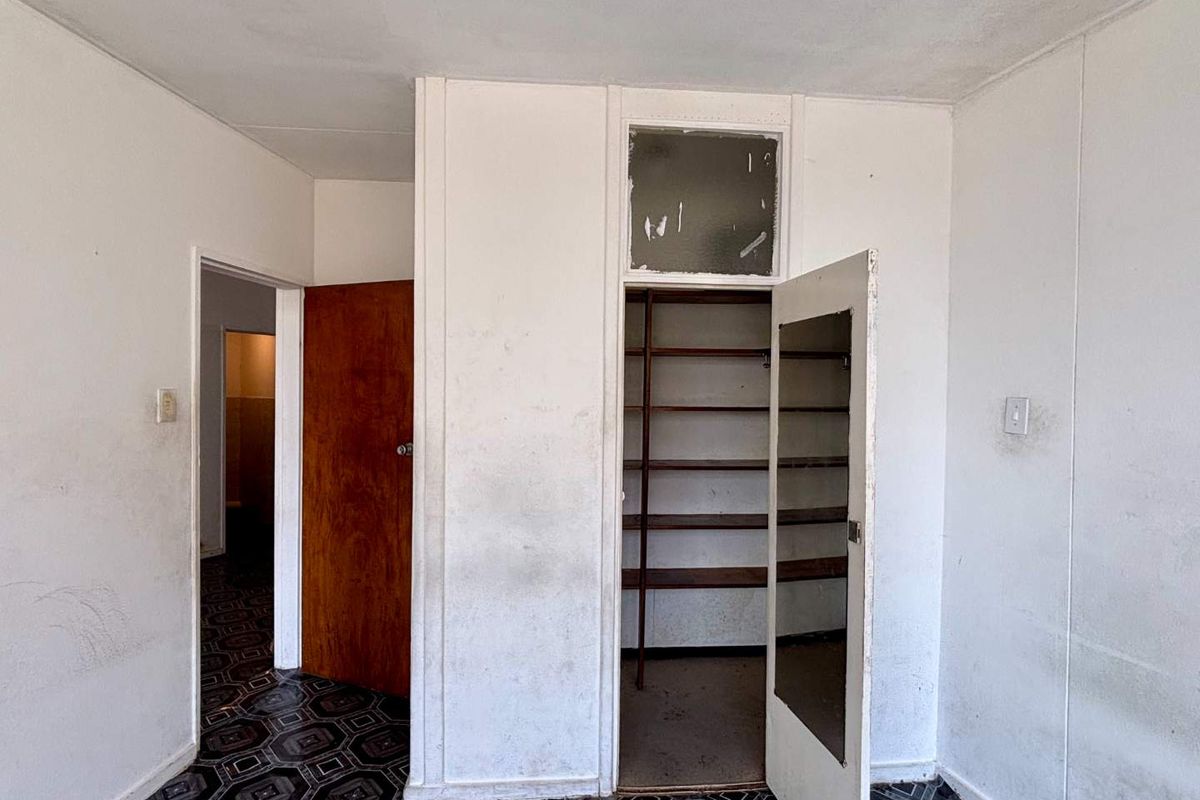 An empty bedroom featuring built-in shelving, a wooden door, and a transom window, requiring cosmetic updates to the walls and flooring.