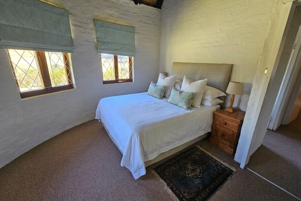 A cozy bedroom featuring rustic white-painted stone walls, exposed thatch ceiling, and two windows with Roman blinds, creating a warm and inviting atmosphere.