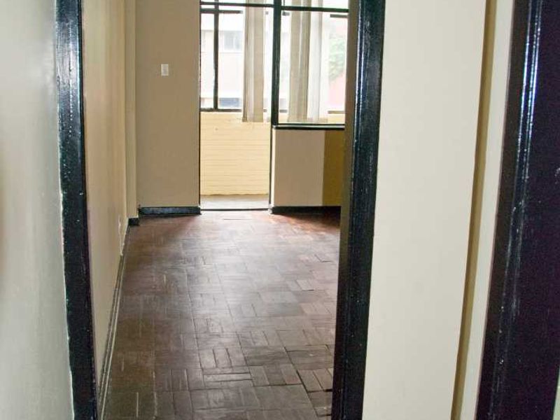 An empty living area featuring original parquet flooring and dark-painted door frames, leading to a bright windowed space.