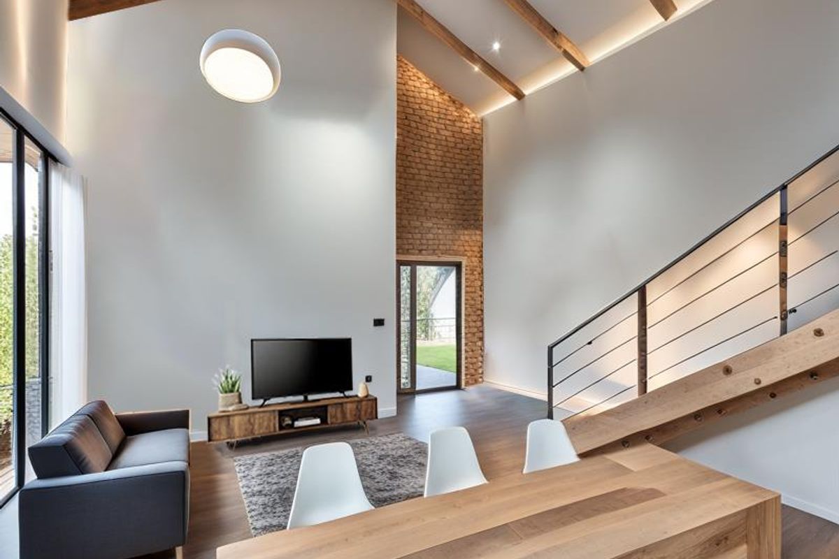 Spacious living area featuring high vaulted ceilings with exposed timber beams, a striking brick feature wall, and a modern staircase with metal balustrades.