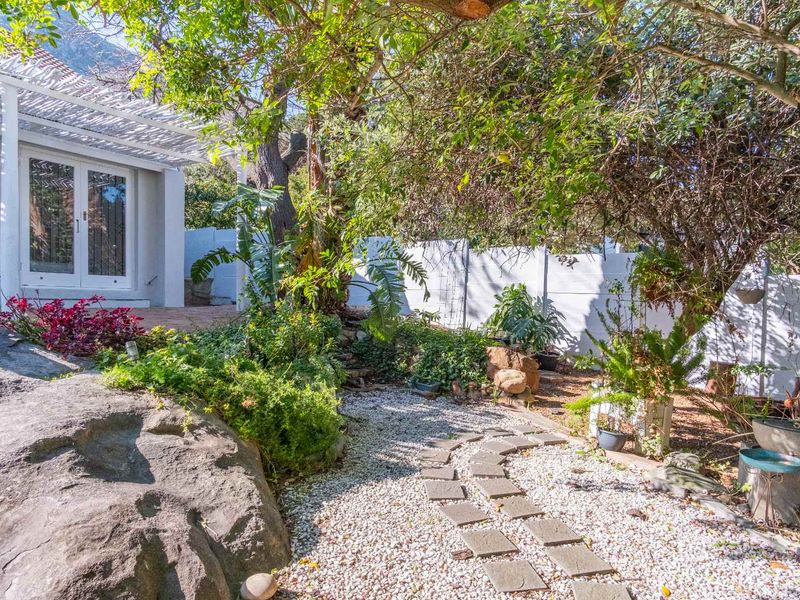 A landscaped garden area featuring a stone pathway, mature trees, a large natural rock feature, and a covered patio area with french doors.