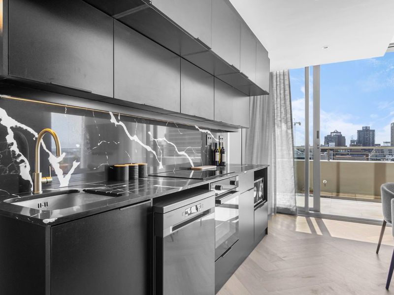 Modern kitchen featuring sleek black cabinetry, marble-look surfaces, integrated appliances, and herringbone flooring, with direct access to a balcony offering city views.