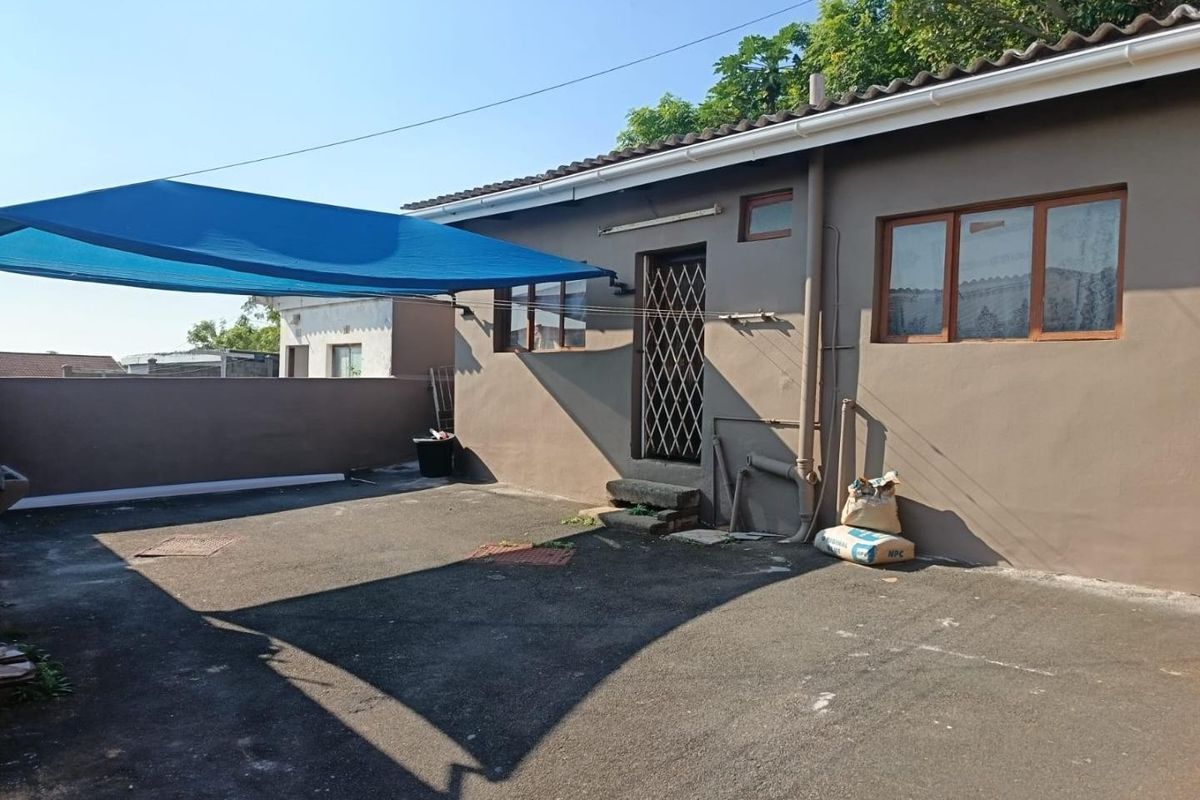 Rear exterior area featuring asphalt paving, a blue shade sail, and a security gate entrance to the main dwelling.