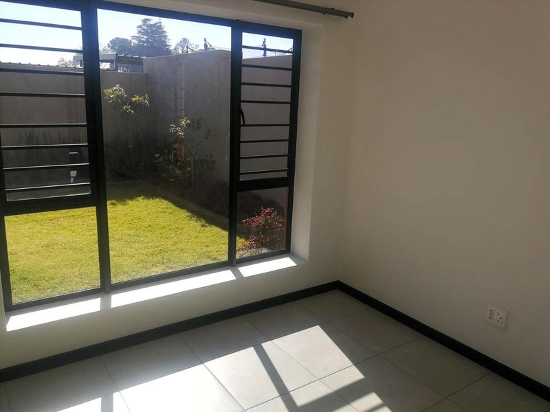 A bright, empty bedroom featuring neutral tiled flooring, black-framed windows with security bars, and a view of the private garden.