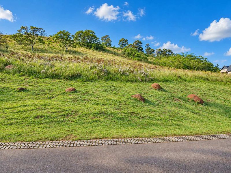 A view of a sloped, grassy residential plot featuring natural rock formations and adjacent paved road access.