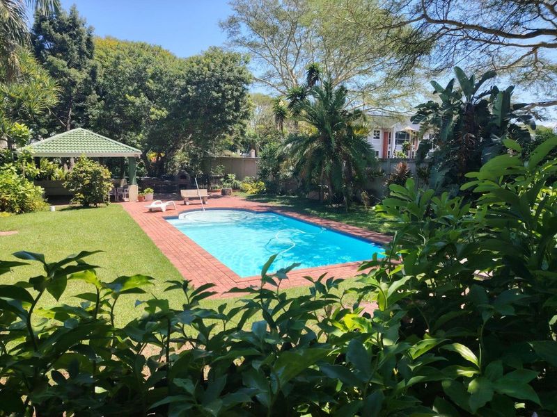 A well-maintained outdoor pool area featuring a swimming pool, brick paved surround, and a covered gazebo set within a landscaped garden.