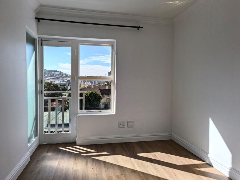 A bright, empty bedroom featuring laminate flooring, crisp white walls, and a glass door leading to a balcony with urban views.