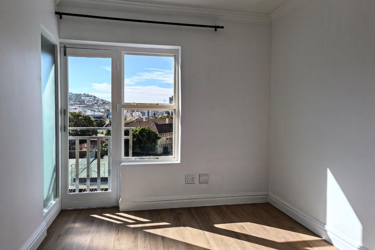 A bright, empty bedroom featuring laminate flooring, crisp white walls, and a glass door leading to a balcony with urban views.