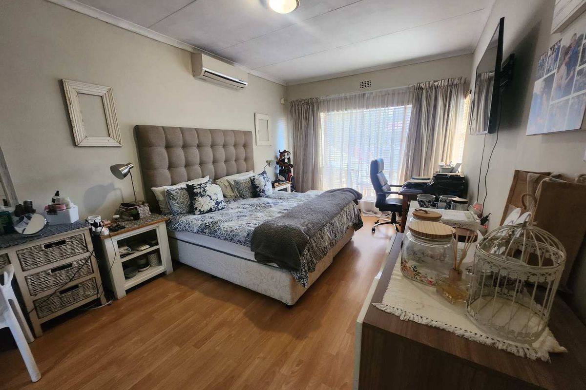 A well-lit bedroom featuring laminate flooring, a wall-mounted air conditioning unit, and large windows with curtains providing natural light.