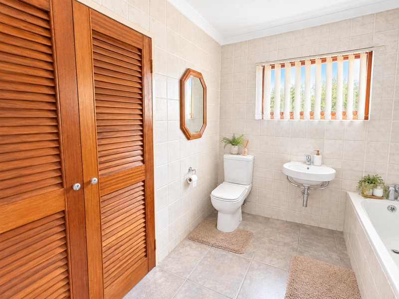 A functional bathroom featuring neutral ceramic tiling, a wall-mounted basin, a close-coupled toilet, a built-in bathtub, and timber louvered storage doors.