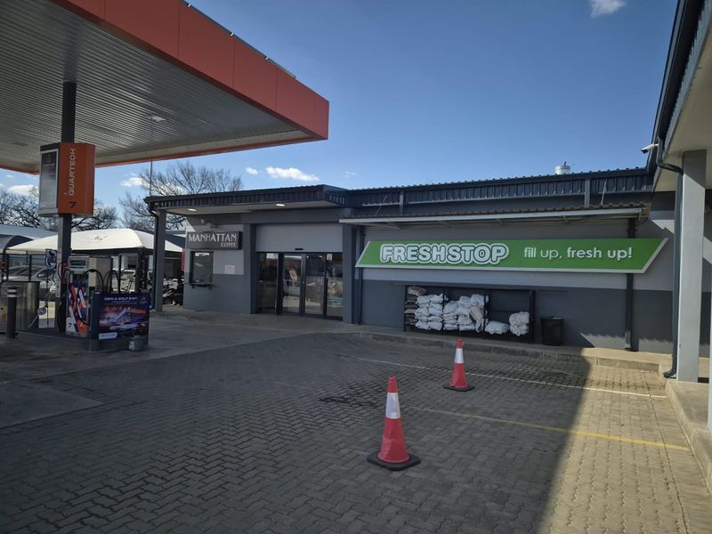 Exterior view of a commercial fuel station forecourt featuring a convenience store entrance, fuel pump canopy, and paved parking area.