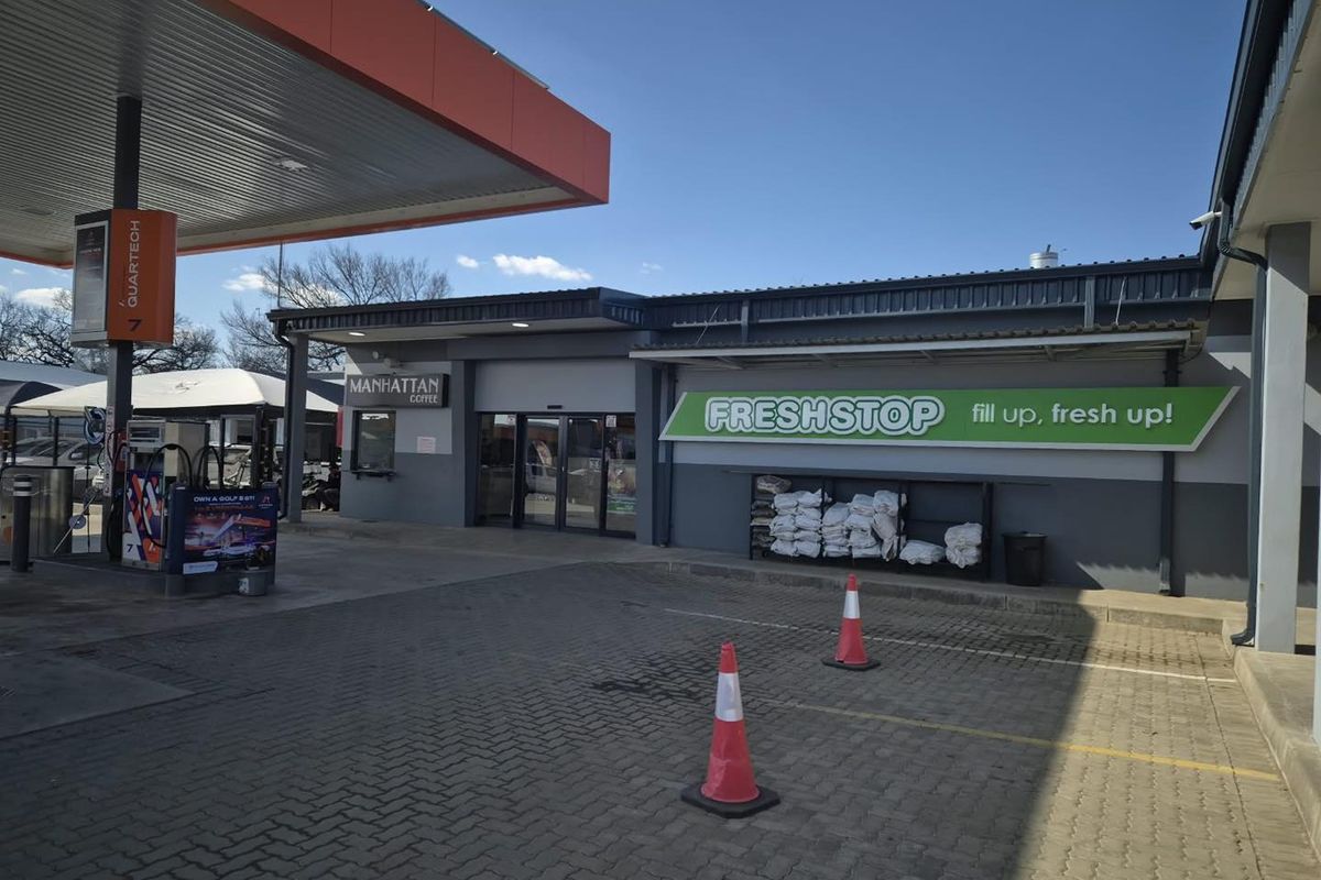 Exterior view of a commercial fuel station forecourt featuring a convenience store entrance, fuel pump canopy, and paved parking area.
