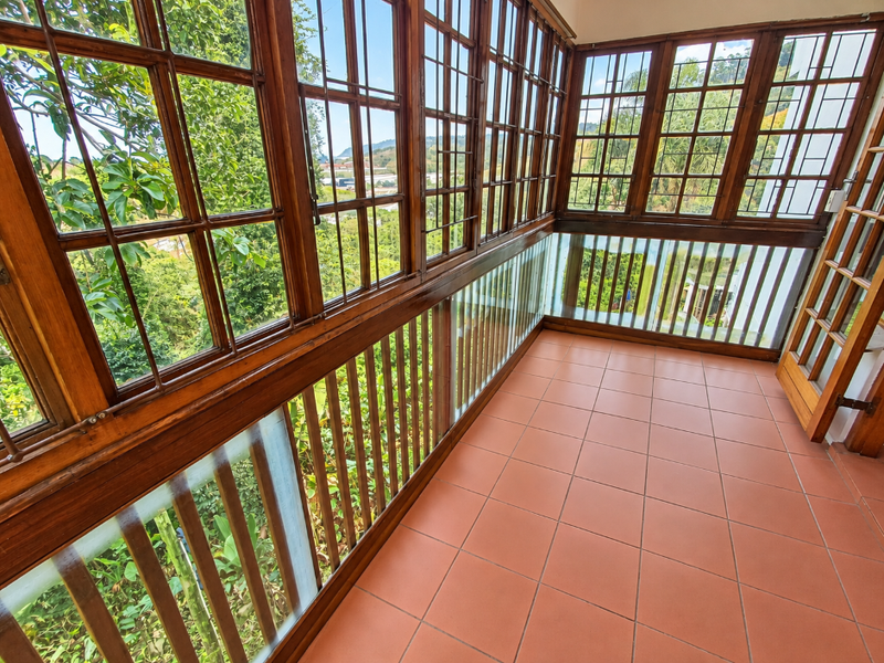 A covered balcony featuring terracotta tiled flooring, extensive wooden-framed windows, and a glass balustrade overlooking the surrounding greenery.