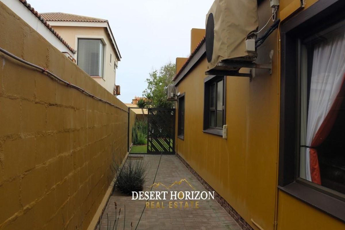 A side passage featuring a paved walkway, boundary wall, and external air conditioning unit, leading towards a rear garden area.