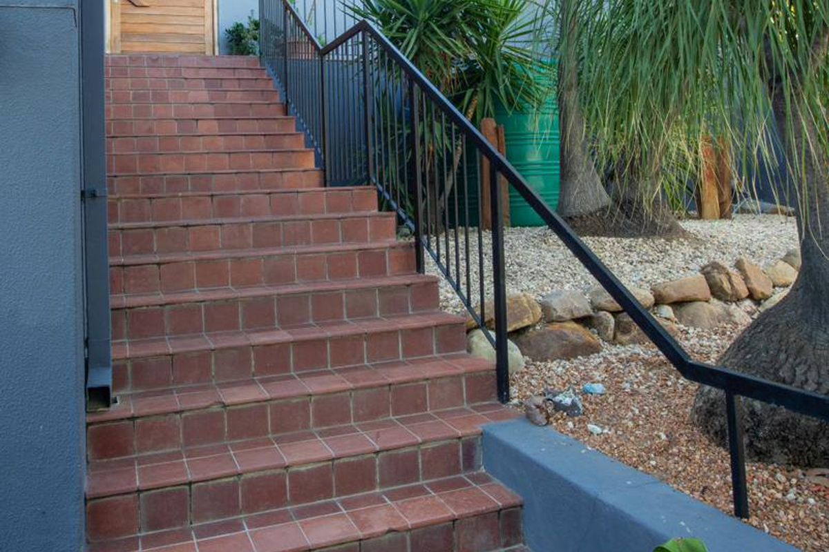 A tiled exterior staircase with a metal handrail leading to a timber entrance door, set against a landscaped garden area.