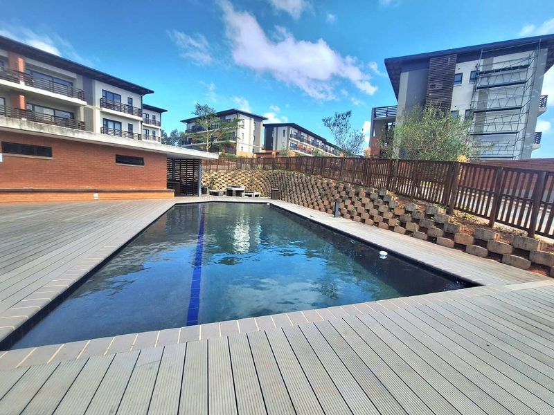 A well-maintained communal swimming pool area featuring timber decking, brick retaining walls, and modern apartment buildings in the background.