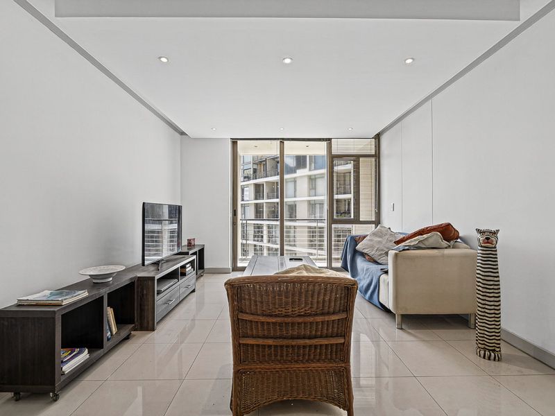 A bright, open-plan living room featuring neutral tiled flooring, recessed ceiling lighting, and large sliding glass doors leading to an outdoor balcony area.