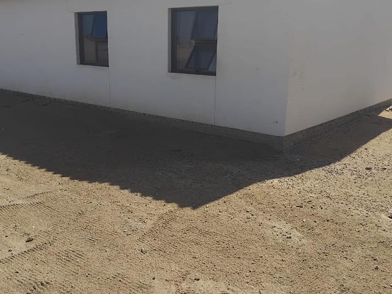 A view of the rear exterior of the house featuring a corrugated roof and casement windows, set against an unlandscaped sandy yard.