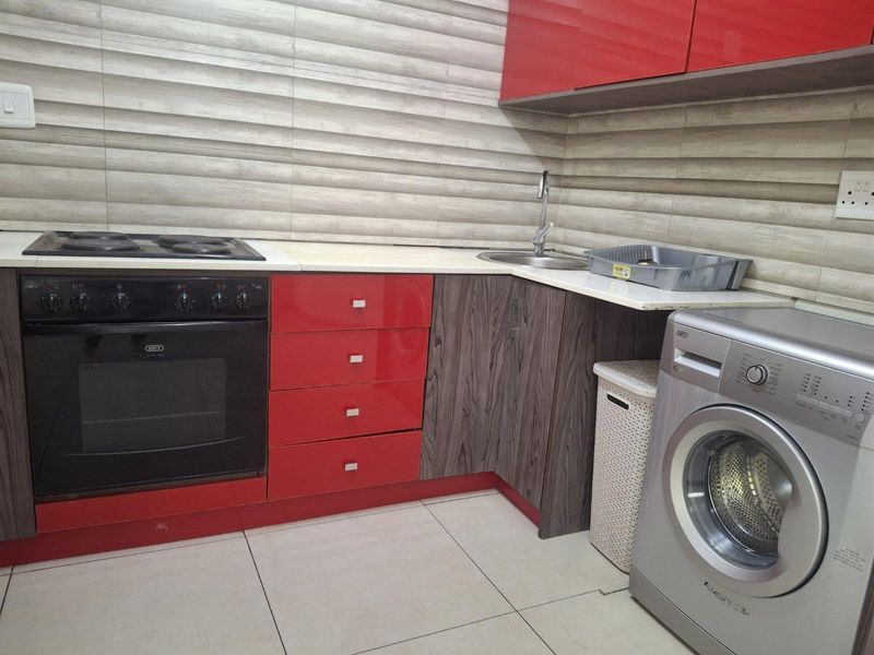 A compact kitchen featuring red and wood-grain cabinetry, a built-in oven with electric hob, stainless steel sink, and modern tiled backsplash.