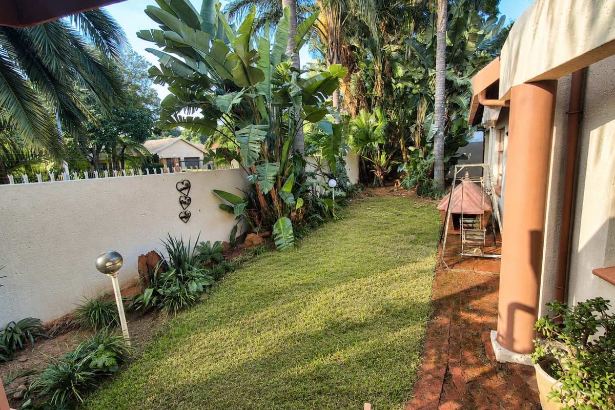 A private rear garden featuring a lawn area, established tropical landscaping, and a covered patio space with brick paving.