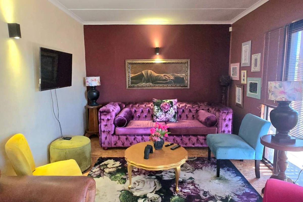 A cozy living room featuring a deep purple accent wall, wall-mounted lighting, and large windows with fitted blinds allowing natural light.