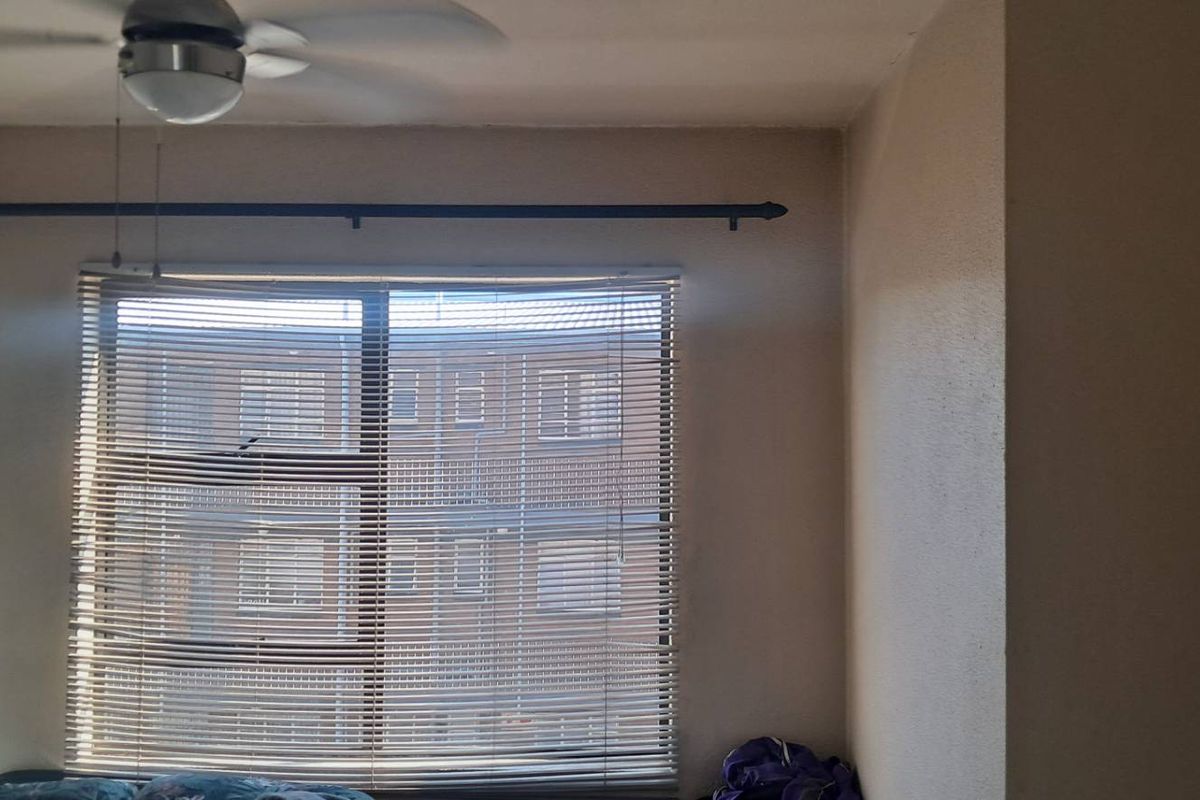A bedroom featuring a ceiling fan and a window fitted with horizontal blinds, offering a view of the neighboring apartment building.