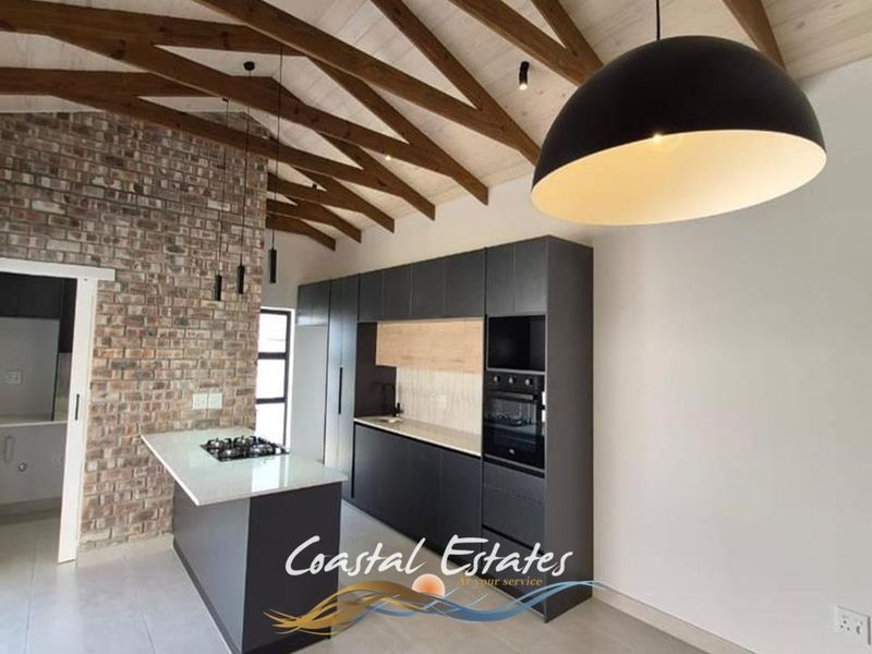 Modern kitchen featuring exposed timber beams, a brick feature wall, sleek dark cabinetry, stone countertops, and a central island with a built-in hob.