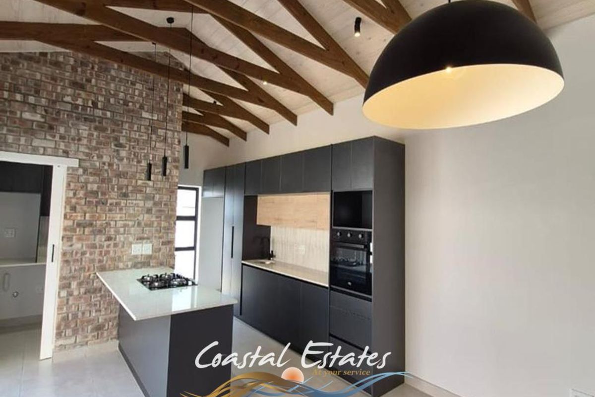 Modern kitchen featuring exposed timber beams, a brick feature wall, sleek dark cabinetry, stone countertops, and a central island with a built-in hob.