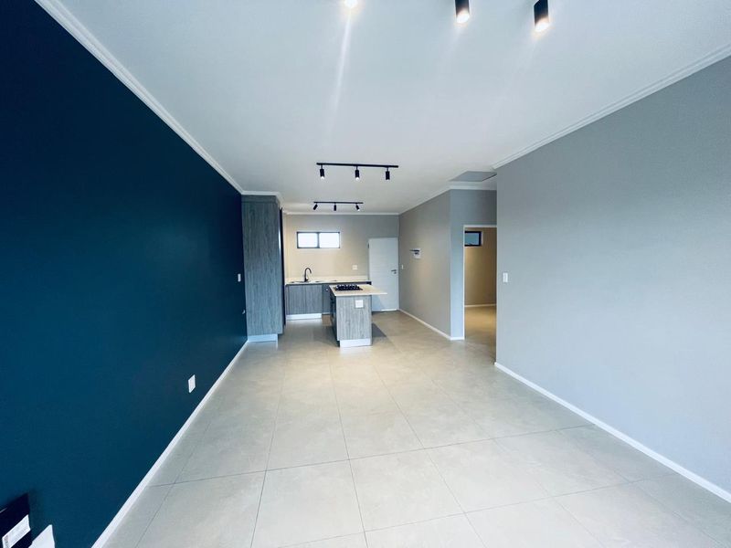 A modern, open-plan living and kitchen area featuring neutral tiled floors, contemporary track lighting, and a functional kitchen island with a built-in hob.