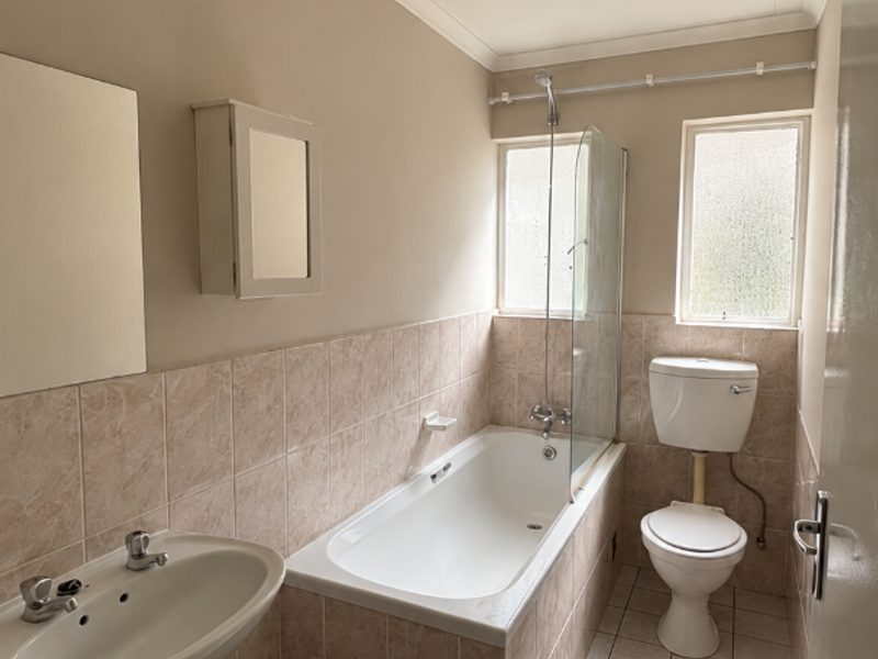 A functional bathroom featuring a bathtub with shower screen, pedestal basin, and toilet, finished with neutral wall and floor tiling.