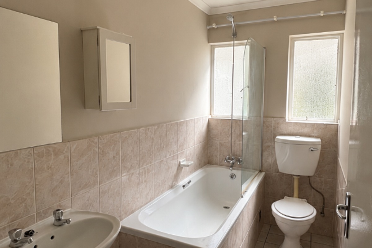 A functional bathroom featuring a bathtub with shower screen, pedestal basin, and toilet, finished with neutral wall and floor tiling.