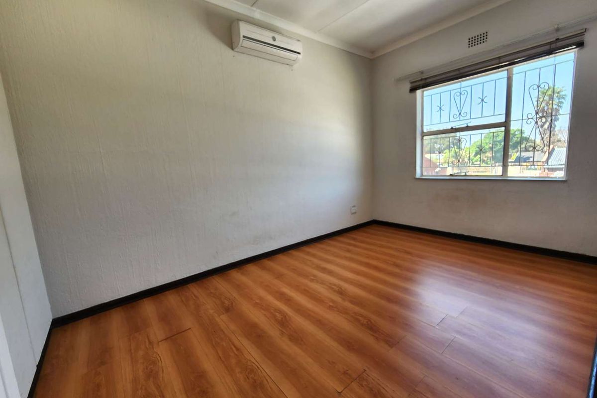 A bright, empty bedroom featuring wood-look laminate flooring, a wall-mounted air conditioning unit, and a window with security bars.