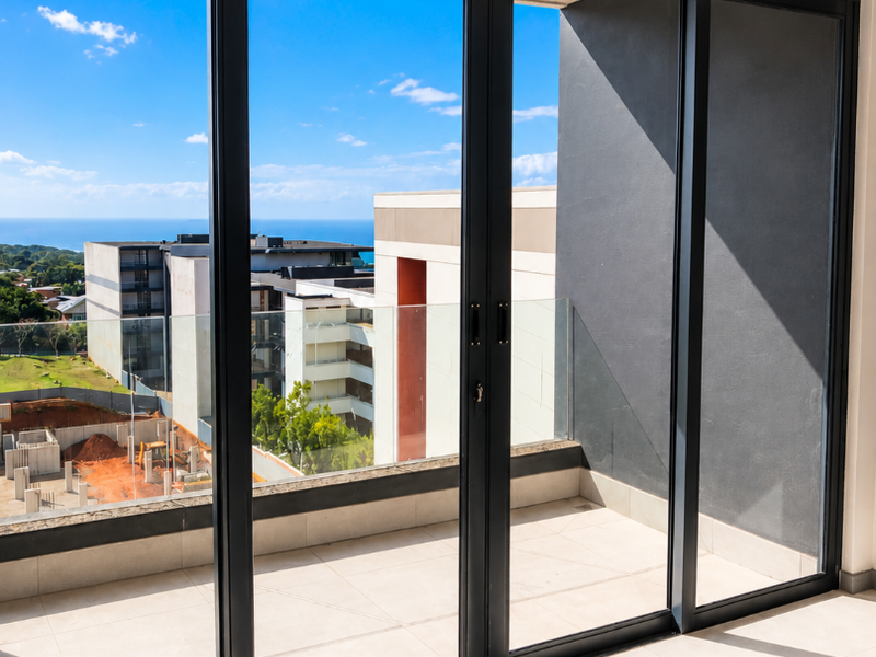 A modern, empty balcony featuring large glass sliding doors, tiled flooring, and a glass balustrade with views of the surrounding area.