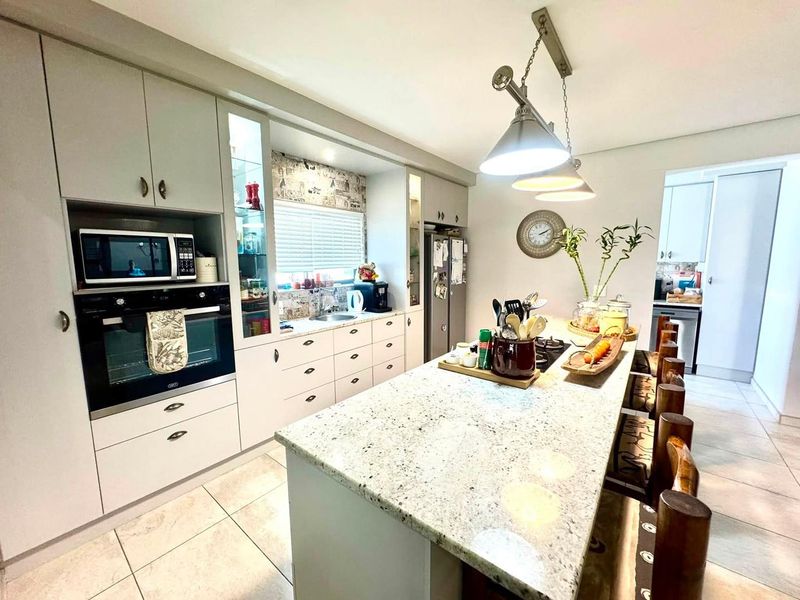 Modern kitchen featuring a large granite-topped island, built-in oven and microwave, ample cabinetry, and tiled flooring, with an adjacent scullery area.