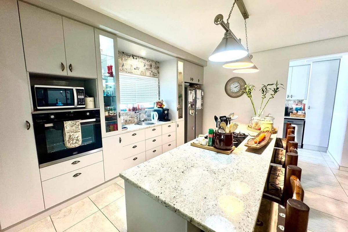 Modern kitchen featuring a large granite-topped island, built-in oven and microwave, ample cabinetry, and tiled flooring, with an adjacent scullery area.