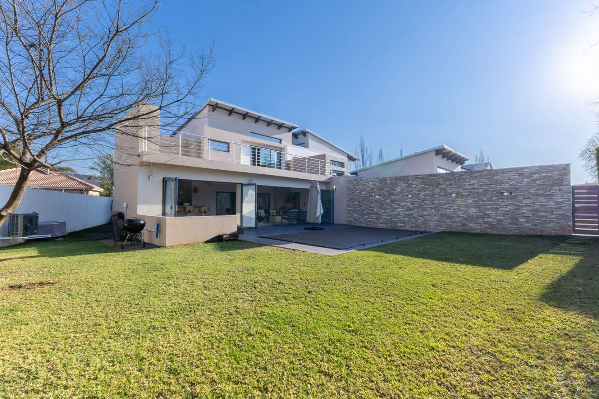 Rear view of a modern two-story house featuring a spacious lawn, stone-clad boundary wall, covered patio, and an upper-level balcony with metal railings.