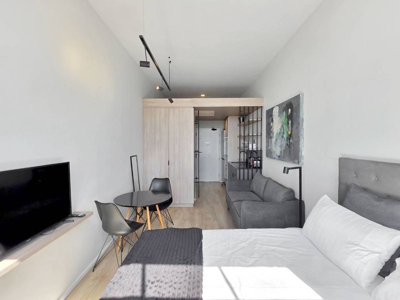 A modern open-plan studio apartment featuring light wood-look flooring, built-in cabinetry, and efficient track lighting in a compact, well-maintained living space.