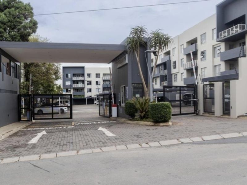 Modern apartment complex entrance featuring a secure gated access point, guard house, and paved driveway with multi-story residential buildings in the background.