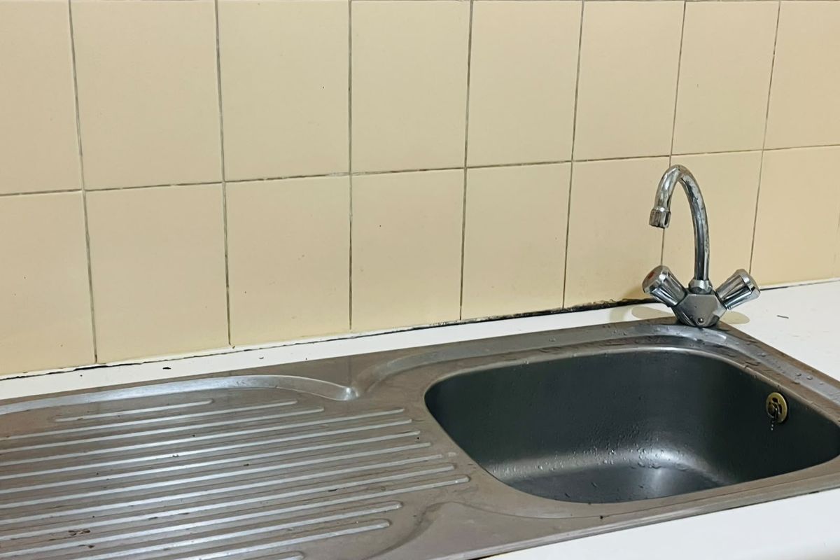 A dated kitchen sink area featuring a stainless steel basin, chrome tap, and beige ceramic wall tiles requiring maintenance.