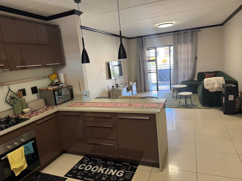 Open-plan living area featuring a modern kitchen with brown cabinetry, stone countertops, built-in oven, and tiled flooring, leading to a balcony.