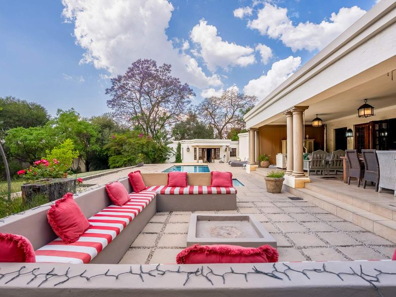 Spacious covered patio featuring built-in concrete seating, a central fire pit, and stone columns, overlooking a private swimming pool and landscaped garden.