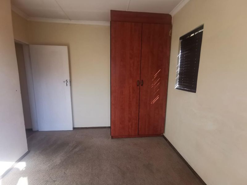 A simple bedroom featuring a built-in wooden wardrobe, carpeted flooring, and a window with fitted blinds.