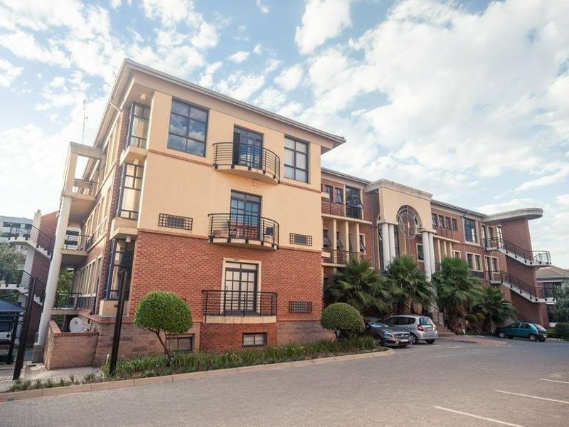 A multi-story office building featuring a brick and plaster facade, external staircases, and private balconies with metal railings, set within a paved parking area.