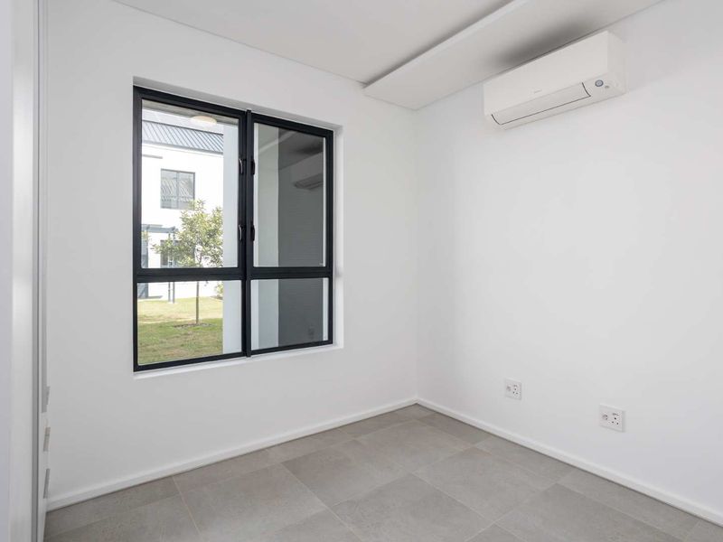 A bright, modern, and empty bedroom featuring neutral tiled flooring, a wall-mounted air conditioning unit, and a large window with black aluminium frames.
