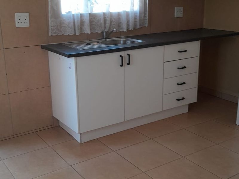 A functional kitchen area featuring white built-in cabinetry, a stainless steel sink, dark laminate countertops, and neutral tiled flooring.