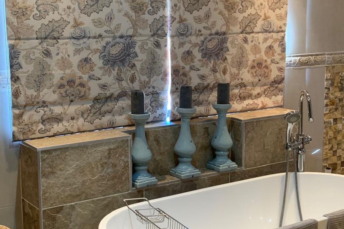 A modern bathroom featuring a freestanding white bathtub with chrome fixtures, complemented by natural stone wall tiling and a decorative Roman blind.
