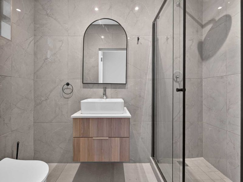 Modern bathroom featuring a wall-mounted wood-finish vanity, vessel sink, arch mirror, and a spacious walk-in shower with black-framed glass enclosure.