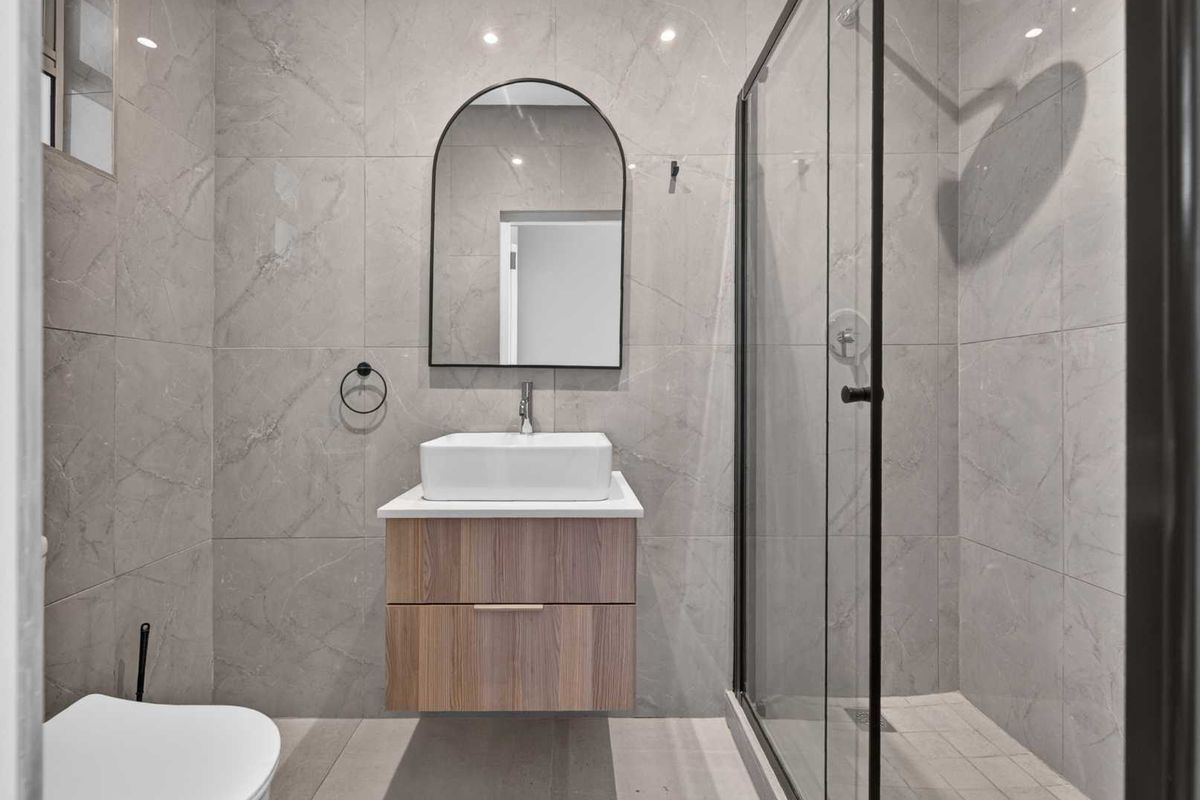 Modern bathroom featuring a wall-mounted wood-finish vanity, vessel sink, arch mirror, and a spacious walk-in shower with black-framed glass enclosure.