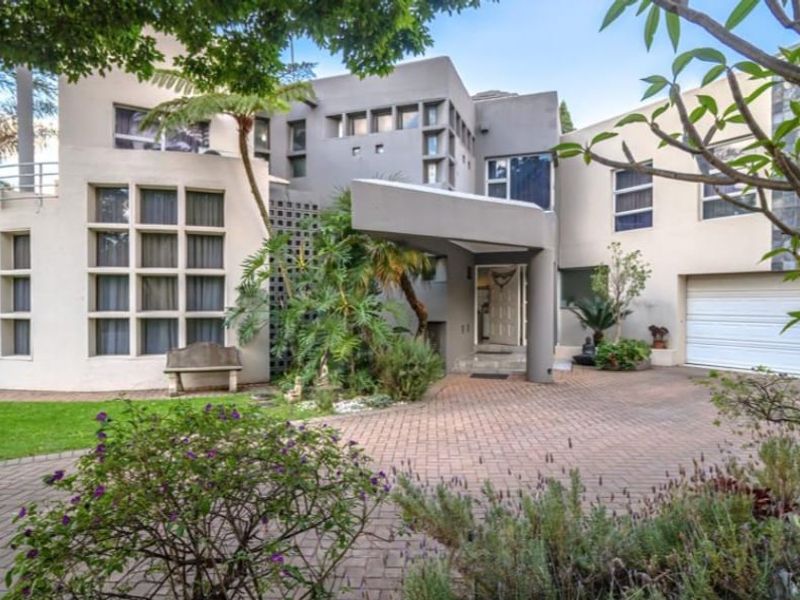 A modern multi-level residential facade featuring a paved driveway, covered entrance, and integrated double garage, set within a landscaped garden.