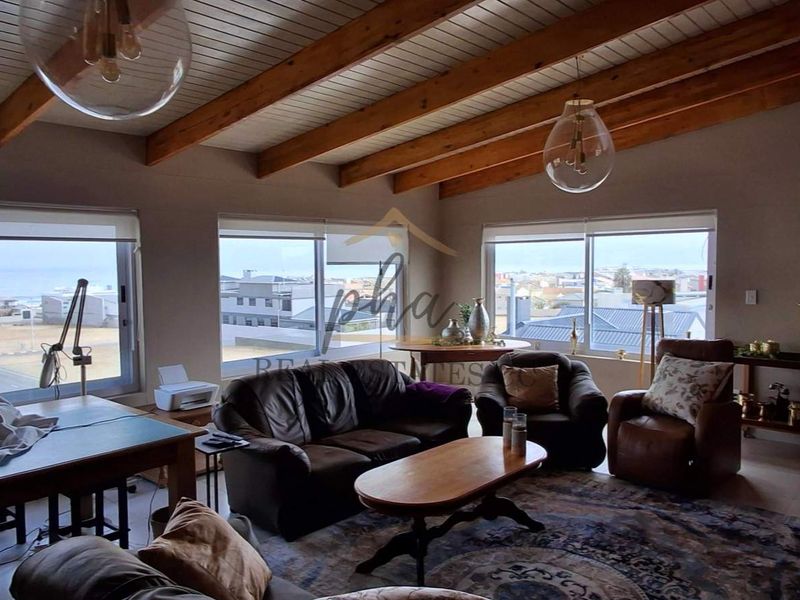Spacious living room featuring exposed timber ceiling beams, large windows with coastal views, and a built-in wood-burning fireplace.